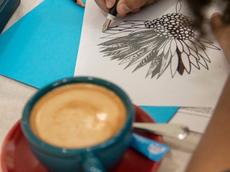 Image of A person draws a detailed flower with a black pen on white paper, while a blue cup of coffee on a red saucer sits nearby on the table. Art supplies and blue paper are also visible.