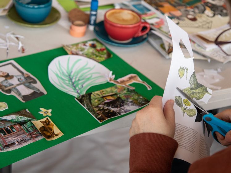Image of A person using blue scissors cuts out images from a sheet of paper for a collage on a green background. The table also holds a red coffee cup, art supplies, and additional collage materials.