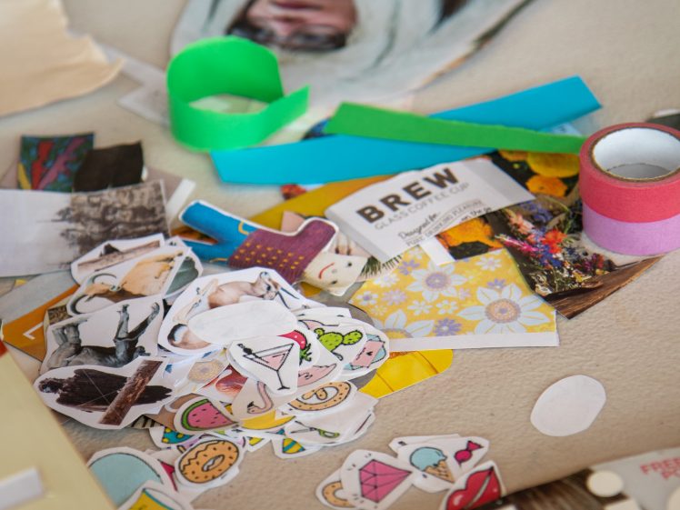 Image of A tabletop scattered with colourful paper cut-outs, stickers of food and drinks, rolls of tape, a green ribbon, and a business card, suggesting a crafting or scrapbooking activity.