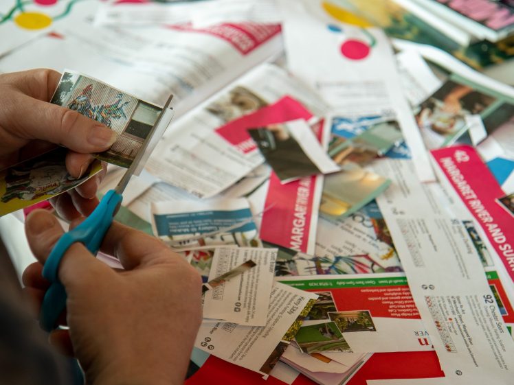 Image of A person uses blue scissors to cut a small photo from a pile of colourful magazine pages and clippings spread out on a table, suggesting arts and crafts or a collage project.