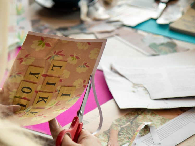 Image of A person uses red scissors to cut a piece of paper with the words I LIKE YOU among floral designs, surrounded by scattered papers, a blue mug, and crafting supplies on a table.