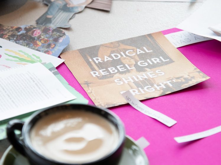 Image of A cluttered desk with craft materials, paper cut-outs, a cappuccino in a black cup, and a card reading “RADICAL REBEL GIRL SHINES BRIGHT.” Scissors and scattered papers are visible in the background.