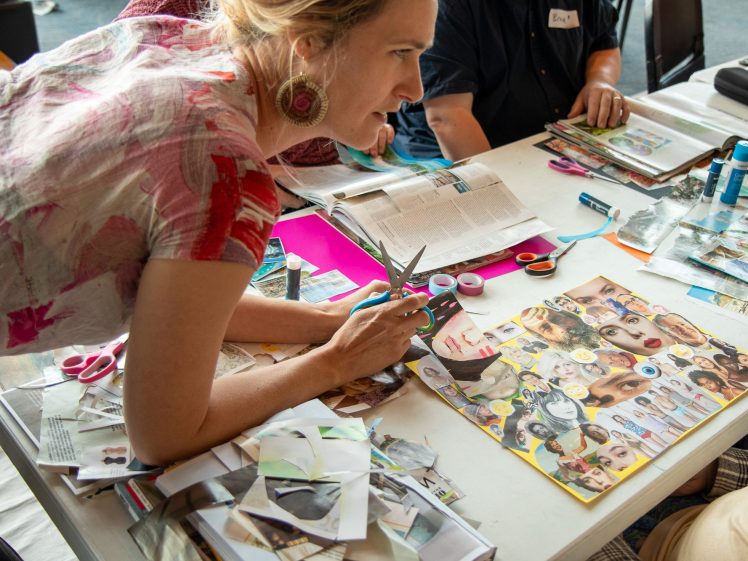 Image of A woman leans over a table covered with magazines, scissors, and art supplies, working on a collage with images of faces whilst others create art beside her.