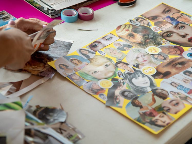 Image of A person cuts magazine clippings at a table, next to a colourful collage of various faces and eyes on yellow paper. Art supplies, including washi tape and scattered paper pieces, are visible on the tabletop.
