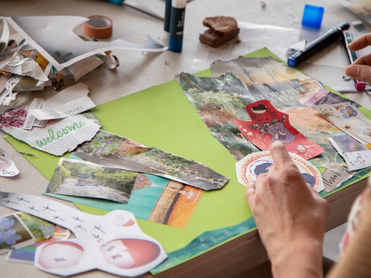 Image of A person arranges colourful magazine cut-outs and stickers on a green sheet of paper, creating a collage. Scissors, glue sticks, pens, and paper scraps are scattered on a white table.