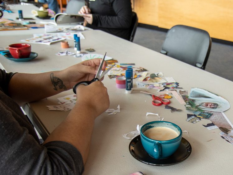 Image of A person cuts out paper pieces for a collage at a table scattered with art supplies, mugs, and coffee. Other people work on similar projects in the background.