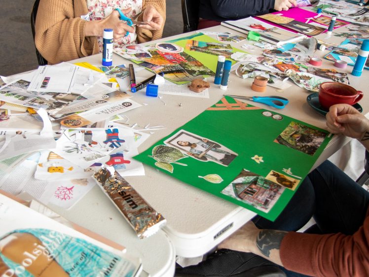 Image of Several people sit around a table covered with magazines, scissors, glue sticks, and collage materials, working on colourful vision boards. One person holds a glue stick whilst another arranges cut-out images on green paper.