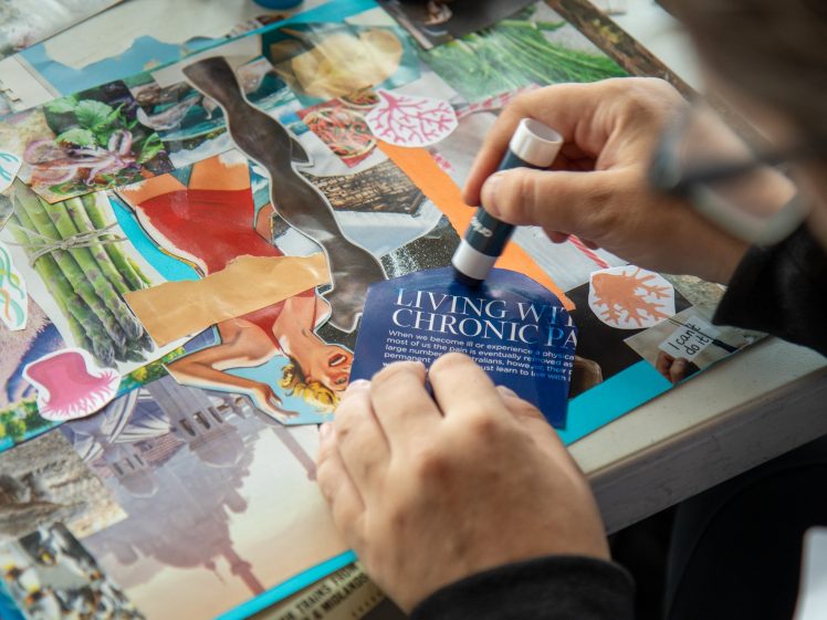 Image of A person uses a glue stick to attach a piece of paper reading “LIVING WITH CHRONIC PA” to a colourful collage of magazine clippings on a table.