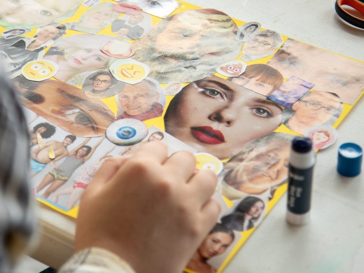 Image of A person’s hand works on a collage of various magazine cut-outs, including faces, eyes, and emojis, arranged on a yellow background. Glue sticks and scissors are nearby on the table.