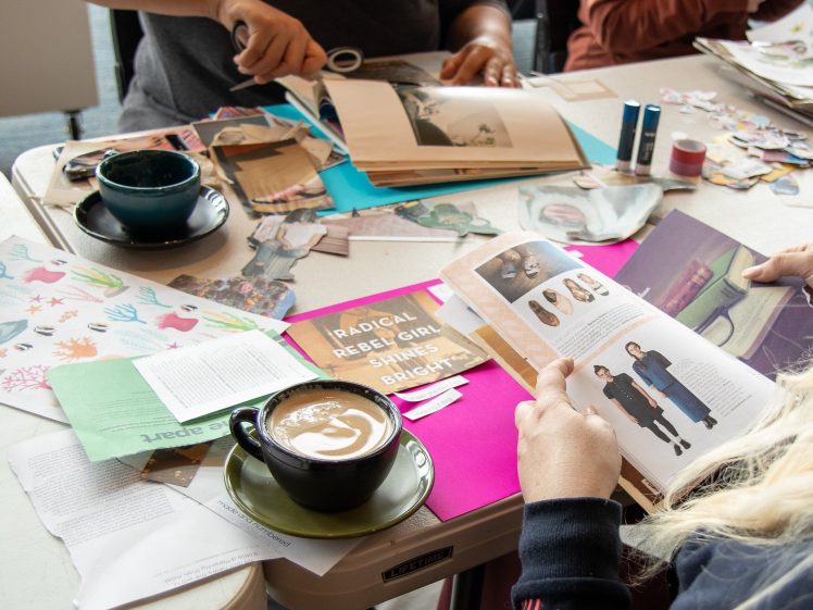 Image of People working on a collage or craft project at a table covered with magazines, papers, scissors, and glue. A latte in a black cup sits nearby, and one person is reading a fashion magazine.
