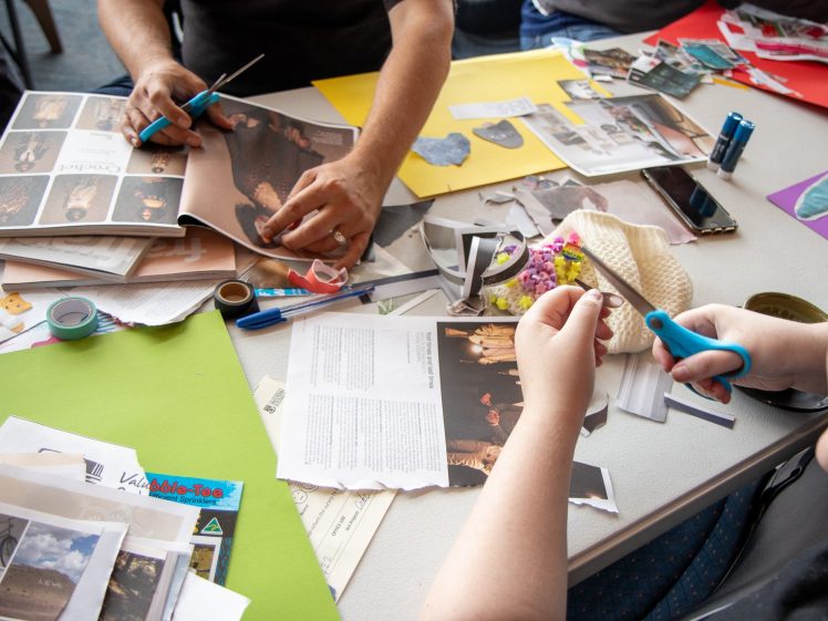 Image of Two people sit at a table covered with magazines, papers, scissors, tape, and craft supplies, working on a collage or scrapbooking project with colourful materials.