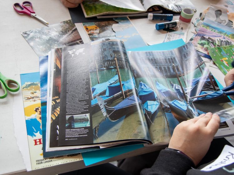 Image of Two people sit at a table doing collage art, with open magazines, scissors, glue sticks, and paper. One person holds a magazine showing gondolas on a canal, whilst cut-outs and craft supplies are spread around.