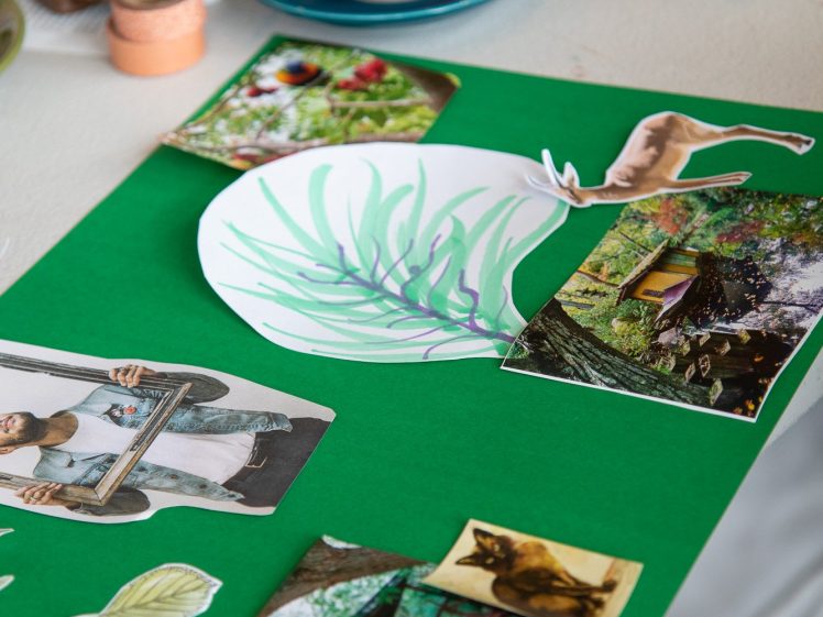Image of A collage in progress on green paper features cut-out images of nature, animals, and people, with art supplies, a cup of coffee, and magazines scattered on the table in the background.