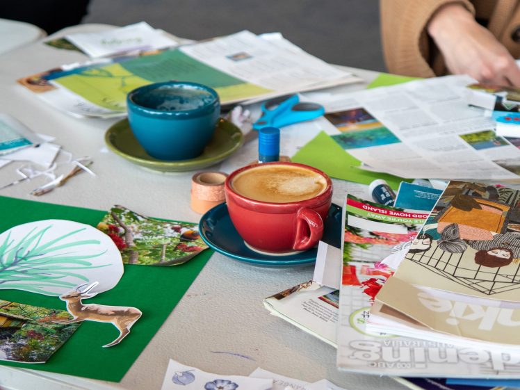 Image of A table with craft supplies, cut-out magazine images, scissors, tape, a blue cup with paint, and a red cup with cappuccino. Two people’s arms are visible as they work on a collage project.