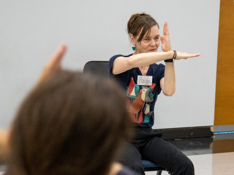 Image of A woman wearing a shirt with a dog print and a name badge reading Paige sits and gestures with her arms extended, palms apart, in front of a blank wall and a wooden door. Another person’s raised arm is visible in the foreground.