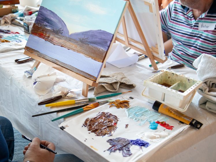 Image of A painting class scene shows people working on landscape paintings of mountains and water, with paint palettes, brushes, and art materials on a table covered with a white cloth.