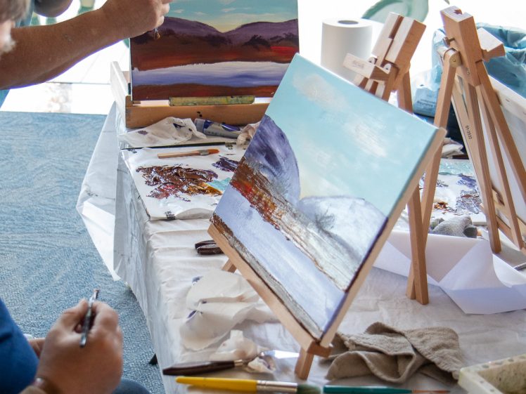 Image of Two people sit at a table painting landscapes on canvas. Palettes with mixed paints and paintbrushes are on the table, and finished and in-progress nature scenes with mountains and water are visible on easels.
