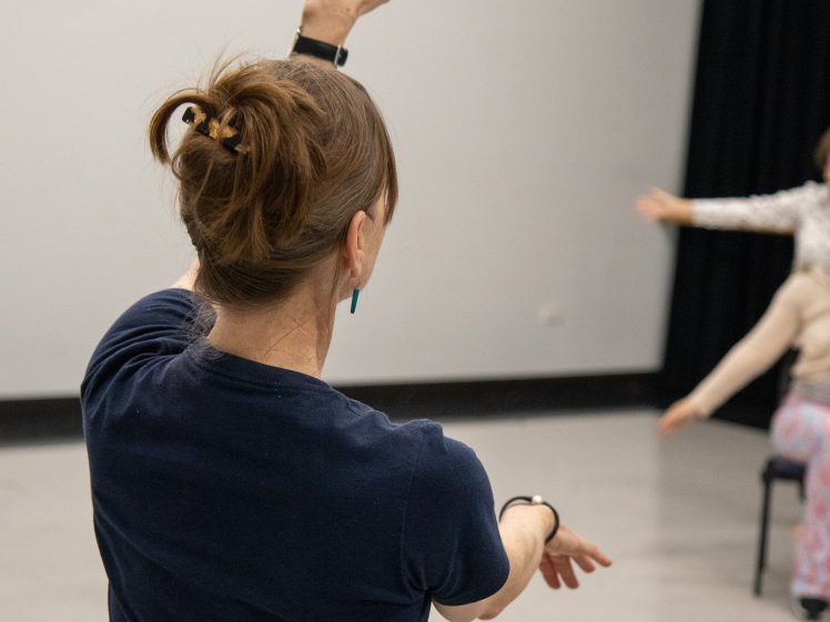Image of A woman with brown hair in a ponytail raises one arm and extends the other whilst participating in a dance or movement class. Another person is visible in the background, also extending an arm.