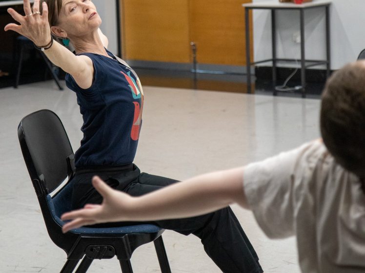 Image of A woman sits on a chair with her arms outstretched in a dance pose, looking upwards. Another person in the foreground mimics her gesture. The setting appears to be a dance studio or classroom.