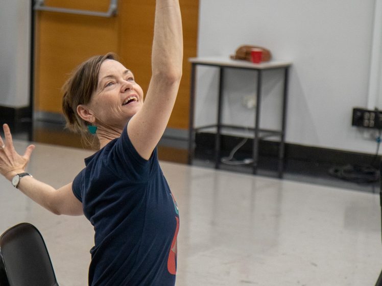 Image of A woman sitting on a chair enthusiastically raises one arm and gestures with the other, smiling and looking upwards in a lively, expressive pose. She is indoors in a room with a table and door in the background.