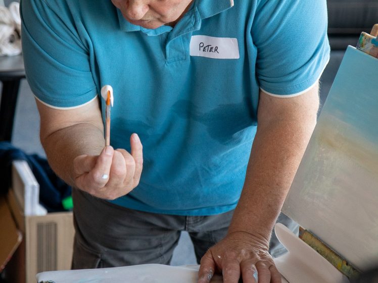 Image of An older man in a blue shirt labelled Peter examines a paintbrush in his hand while standing at a table with paint supplies and a canvas in front of him.