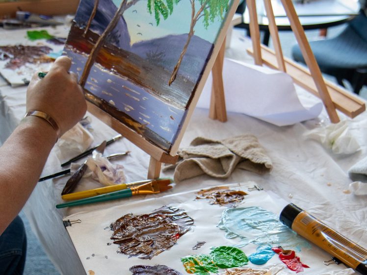Image of A person paints a landscape of trees, water, and mountains on a canvas at an easel. A palette with various paint colours and brushes sits on the table nearby.