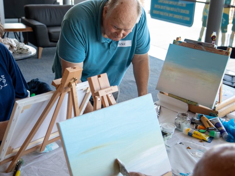 Image of An older man in a blue shirt leans over a table, watching someone paint a landscape on a canvas. Art supplies and two easels are on the table. Another person’s arm is visible, painting on a canvas in the foreground.