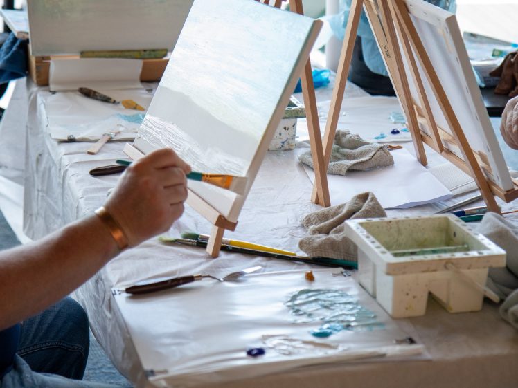 Image of People painting on canvases set on easels at a table covered with art supplies, paint, and brushes. The focus is on hands working on art projects in a creative setting.