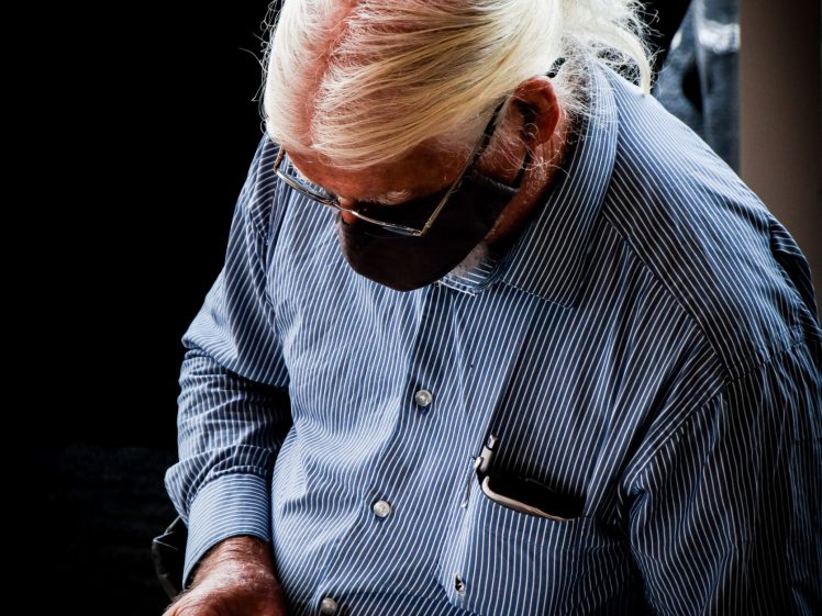 Image of An elderly person with long white hair and glasses, wearing a striped shirt and face mask, is reading a booklet. A book and a bottle are on a nearby table.