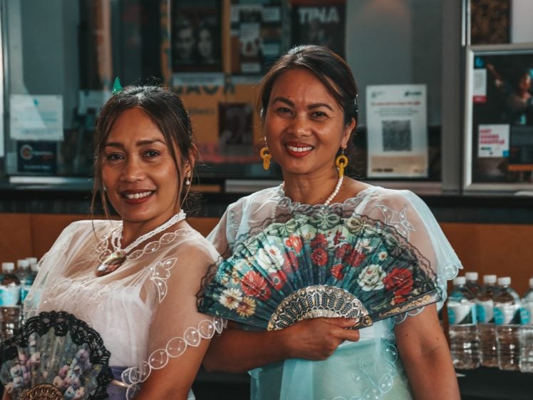 Image of Two women in traditional attire smile while holding ornate handheld fans, standing indoors in front of a table with water bottles and display boards in the background.