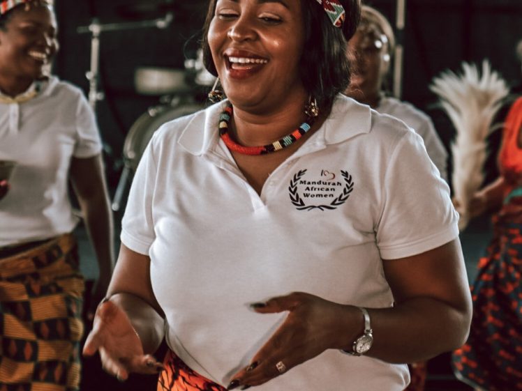Image of A smiling woman wearing a beaded headband and necklace balances a wooden bowl on her head. She is dressed in a white polo shirt and a brightly patterned orange wrap skirt, dancing with others in traditional attire in the background.