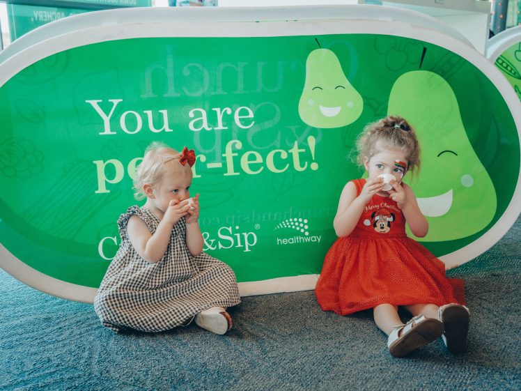 Image of Two young girls sit on the floor in front of a green sign that says “You are pear-fect!” Both are drinking from small cups and wearing dresses, one in checked black and white and the other in red.
