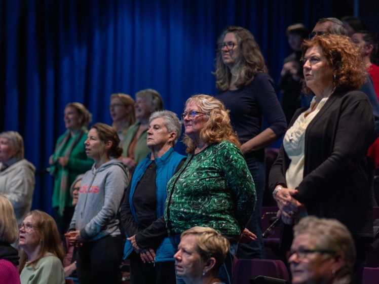 Image of A group of people, mostly women, stand attentively in tiered seating in a dimly lit auditorium with dark blue curtains in the background.