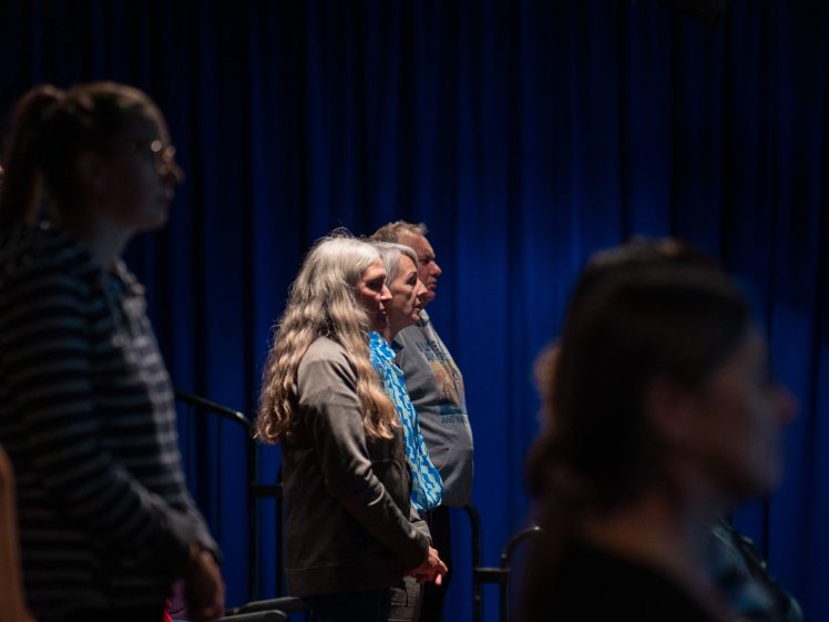 Image of A group of people stand quietly in a dimly lit room with blue curtains, appearing attentive and focused on something out of the frame.