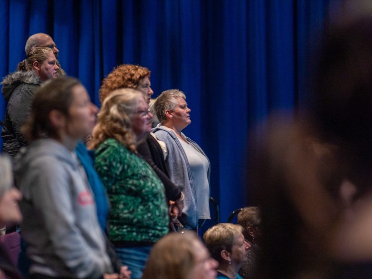 Image of A group of people stand closely together in an audience, facing forward with attentive expressions, against a backdrop of deep blue curtains.