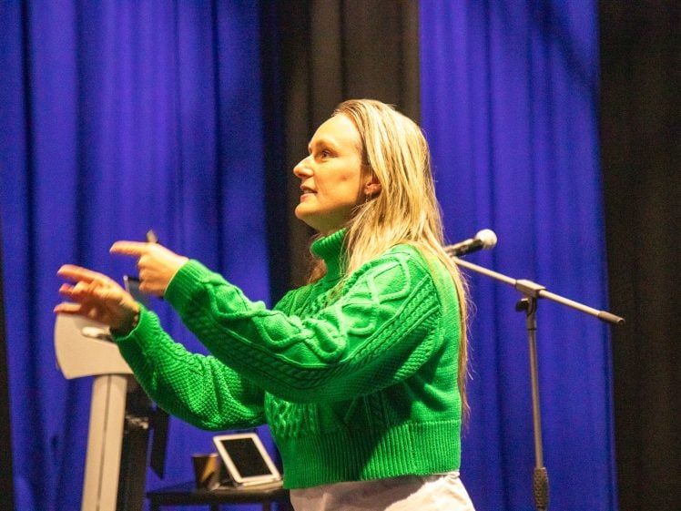 Image of A person wearing a bright green jumper gestures with both hands on stage, with a microphone stand and blue-lit curtains in the background.