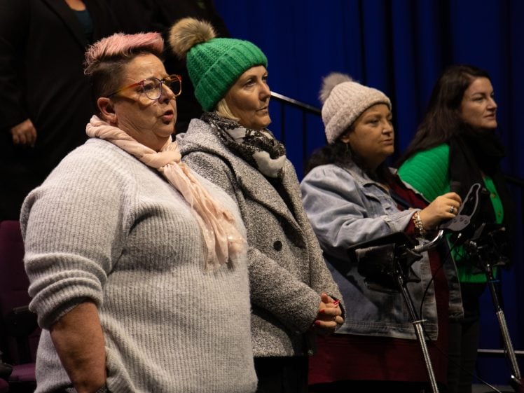 Image of Four women stand indoors, facing forwards. Three wear knitted hats and winter clothing, one wears glasses and a scarf. Two women stand behind mobility aids. The background is dark blue with theatre seating visible.