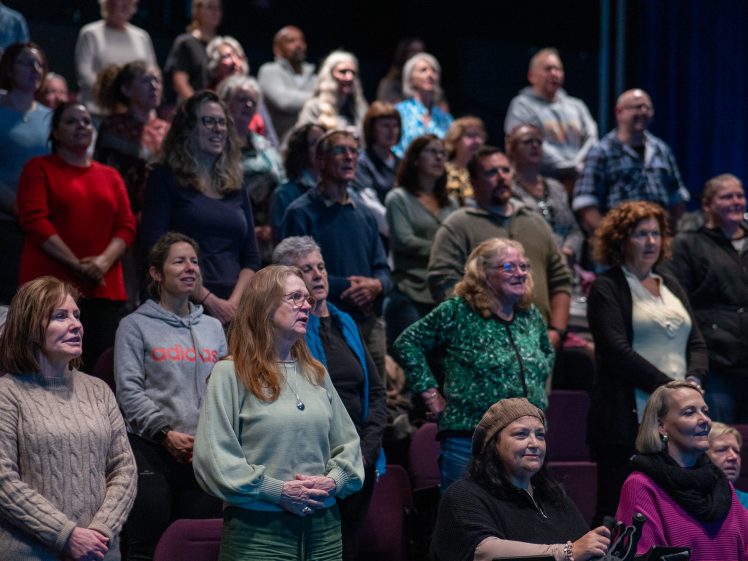 Image of A diverse group of people of various ages standing and singing together in a dimly lit auditorium, some smiling and all appearing engaged in the activity.