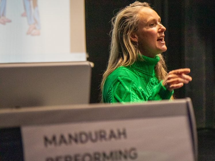 Image of A person in a bright green jumper speaks passionately in front of a projector screen, with a sign reading Mandurah Performing Arts Centre visible in the foreground.