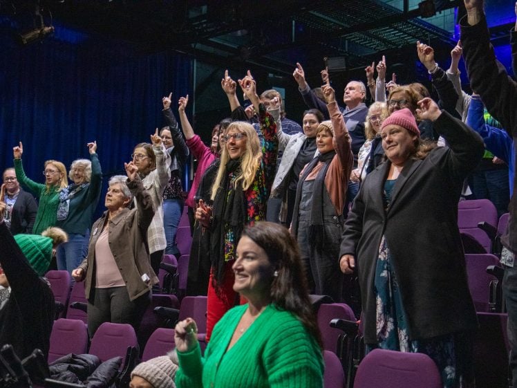 Image of A group of people standing in theatre seats, smiling and raising one finger in the air, appearing joyful and engaged, with purple chairs and dark curtains in the background.