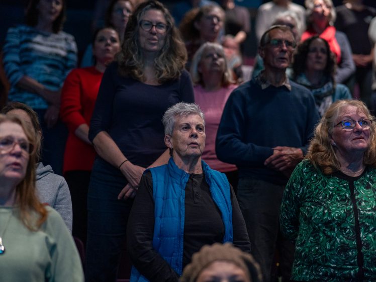 Image of A group of older adults, both women and men, stand closely together in an auditorium, attentively looking forward. Most have neutral expressions and are dressed in casual, warm clothing.
