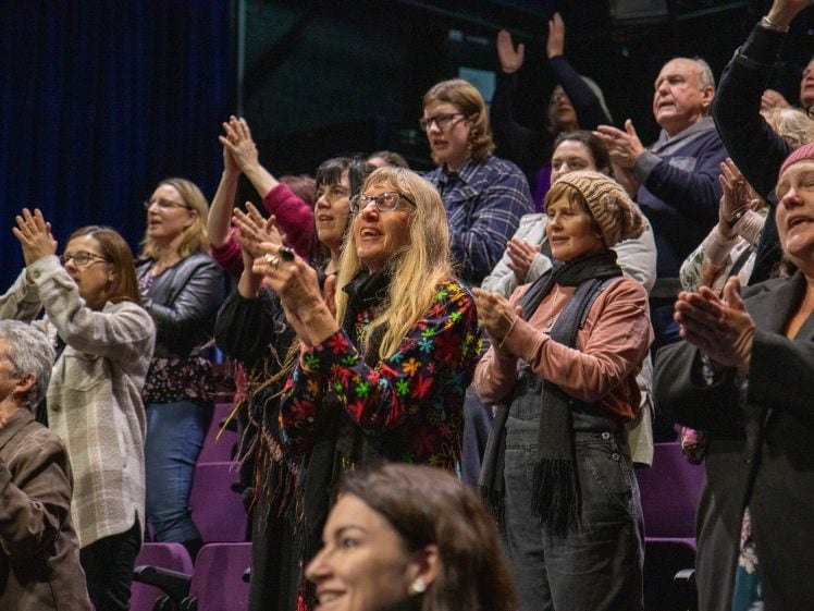 Image of A diverse group of people stand and applaud enthusiastically in a theatre, some smiling and cheering, dressed in casual winter clothing.