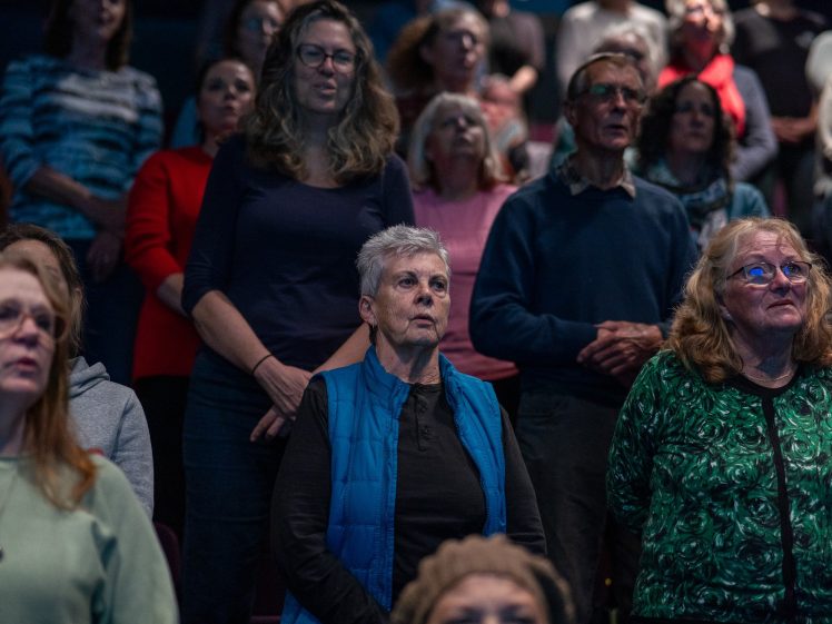 Image of A group of people of various ages stand closely together indoors, appearing attentive and focused, as if listening to someone or something off-camera. The lighting is dim, suggesting an evening event or performance.