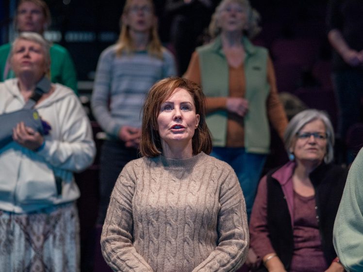 Image of A woman in a beige jumper stands in the foreground, looking ahead with other adults blurred behind her, all appearing engaged in an indoor group setting.