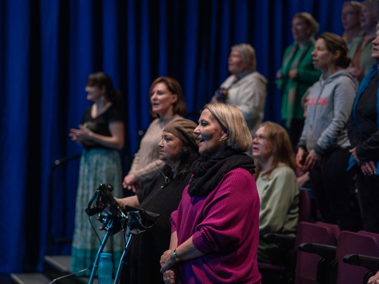 Image of A group of women stand and sing together in a dimly lit theatre, with blue curtains in the background and rows of seating visible.