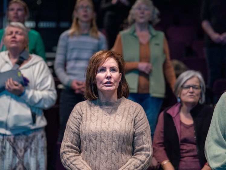 Image of A woman in a tan cable-knit jumper stands among a group of women in a dimly lit room, all facing forwards with serious expressions, possibly participating in a gathering or event.