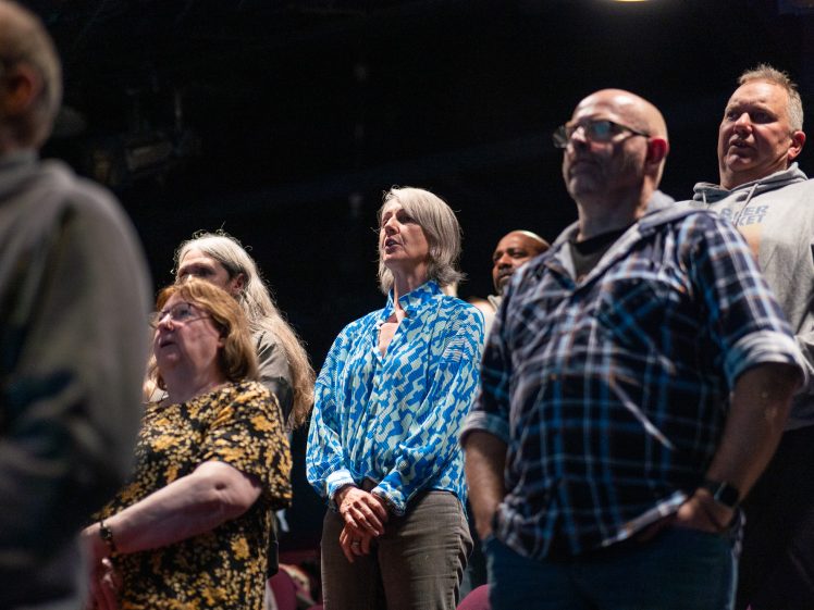 Image of A diverse group of adults stand together in a theatre, attentively facing forward. The setting is dimly lit, and the people appear focused and engaged.