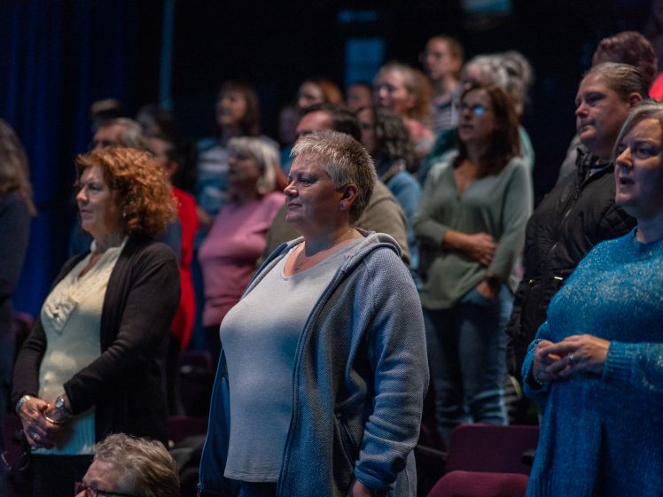 Image of A group of adults, mostly women, stand closely together in an auditorium, appearing attentive and focused on something out of view. The lighting is dim, highlighting the people in the foreground.