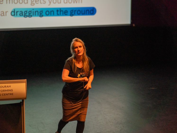 Image of A woman stands on stage speaking, gesturing with her hand. Behind her is a screen with highlighted text and a lectern labelled Mandurah Performing Arts Centre. The stage area is dimly lit.