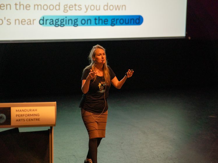 Image of A woman stands on stage speaking into a headset microphone beside a lectern labelled Mandurah Performing Arts Centre. Behind her, a screen displays partially visible song lyrics with highlighted text.